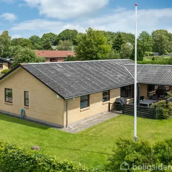 Et gulstenshus har en stor have med græsplæne og terrasse. Flagstangen står foran huset. Landskabet består af omkringliggende træer og nabohuse.