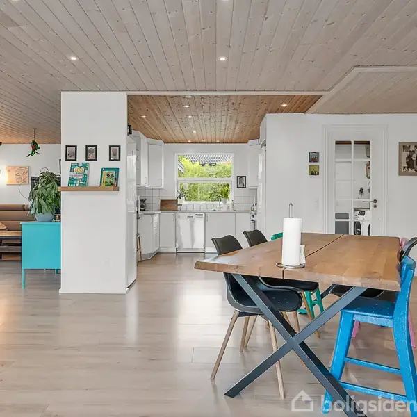 Et moderne spisebord står i forgrunden med farverige stole omkring det. I baggrunden ses en stue med sofa, kunst på væggen og et køkkenområde med hvide skabe.