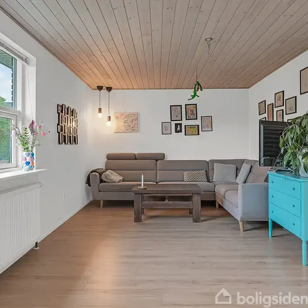 Grå sofa står stille i en lys dagligstue med træloft og gulv. Billeder hænger på væggen. Et blåt skab er til højre, med en stor plante ovenpå.
