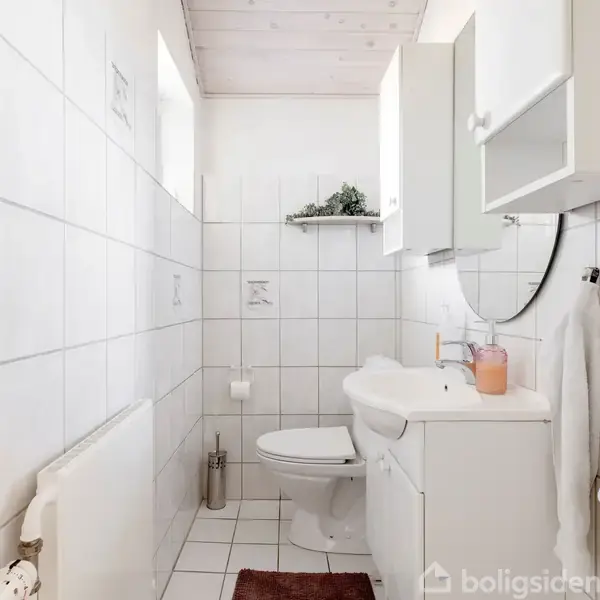 Toilet står ved siden af vask i et lysflisebelagt badeværelse med hvid inventar; håndklæder hænger på væggen, og et lys står ved vasken.