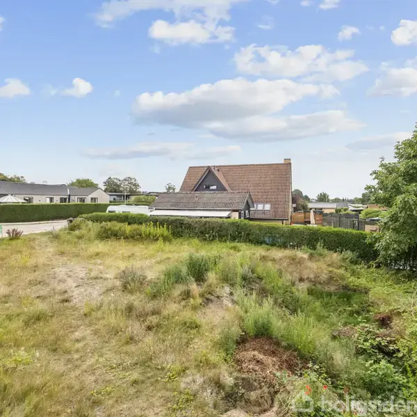 En trekanthus med brunrødt tag står bag en åben græsbeklædt grund. Omgivet af hække og træer under en blå himmel med hvide skyer.