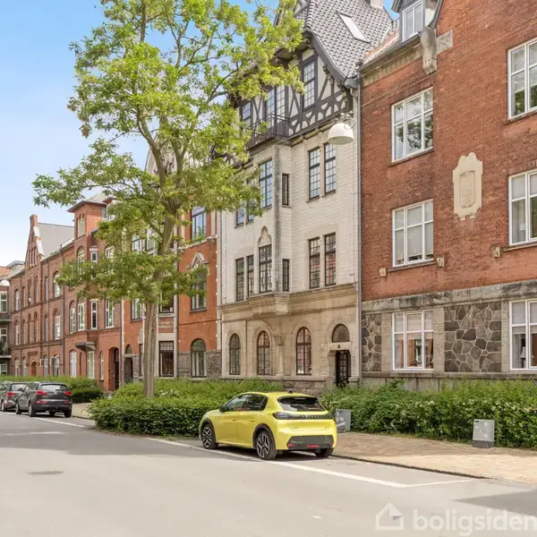 En række ældre murstensbygninger står langs en gade med parkerede biler. En gul bil er parkeret ved kantstenen, mens grønne træer og buske omgiver det hele.