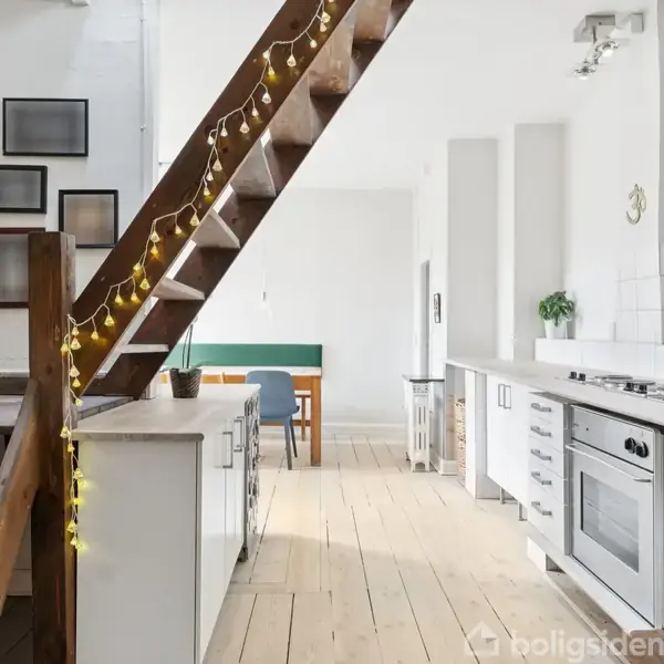 Trappe dekoreret med lyskæde fører opad i et lyst, moderne køkken med hvide fliser, trægulv og bordplade. Spiseområde med grøn bænk ses i baggrunden.