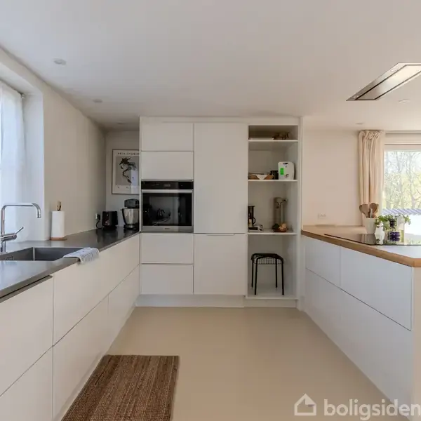 Et moderne køkken med hvide skabe indeholder en indbygget ovn. Sollys strømmer ind gennem to store vinduer, der oplyser rummet med en køkken-ø og vask.