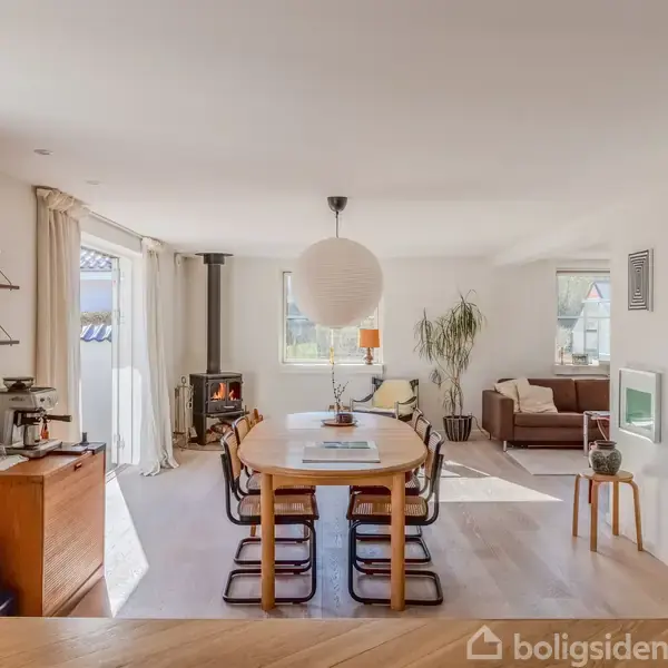 En stue med spisebord til otte personer i centrum, omgivet af sofa, pejs og planter. Store vinduer lader lys ind. Hylder og bord ved siden af tilføjer opbevaring.