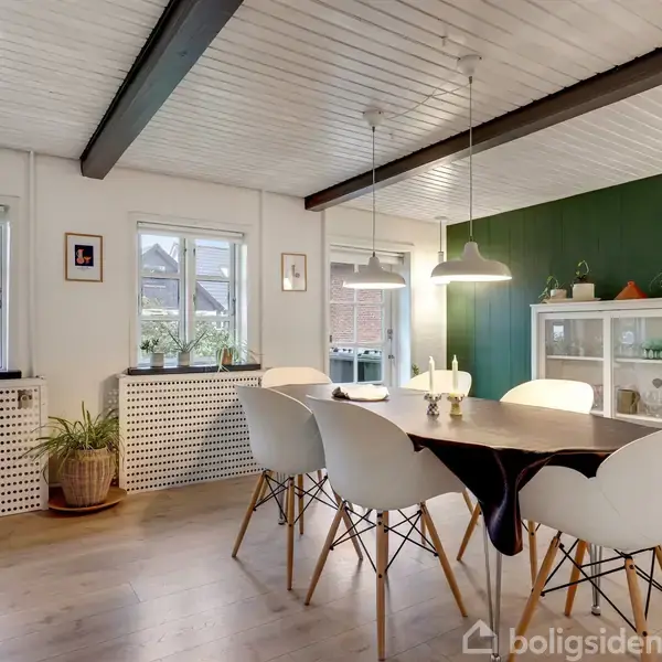 Spisebord omgivet af hvide stole står i et lyst, moderne rum med grønmalet væg. Over bordet hænger lamper. En hvid reol med pyntegenstande ses til højre.
