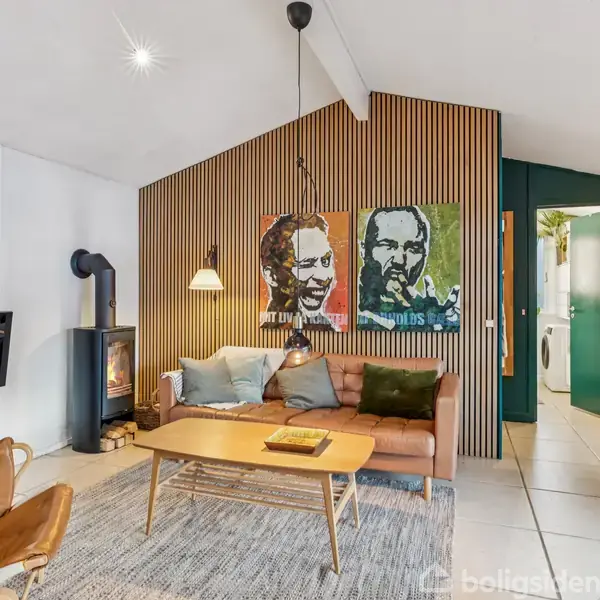 Stue med brun lædersofa, hvorpå der hviler flere puder. Til venstre brændeovn og lænestol. På væggen ses kunstværker. I baggrunden er lysindfald fra vinduer og dør til bryggers.