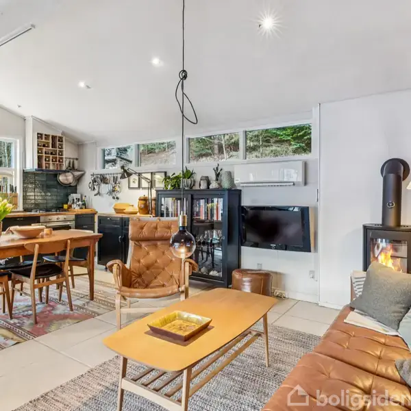 En moderne stue kombineret med et åbent køkken. Indretningen inkluderer sofa, lænestol, spisebord og brændeovn. Store vinduer slipper naturligt lys ind. Møblerne er træfarvede med teksturerede tæpper.