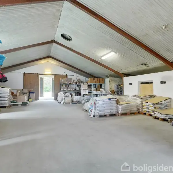 Et stort rum med højt til loftet er fyldt med stabler af sække og nogle farverige tæpper. Rummet har metalhylder og nogle paller, der skaber et lagerlignende miljø.