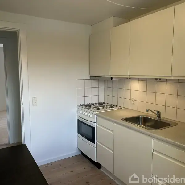 Et køkken med hvide skabe og skuffer, en ovn med kogeplade og en stålvask. Flisevæggen bag vasken giver rummet et rent udtryk. En dør fører til et andet rum.