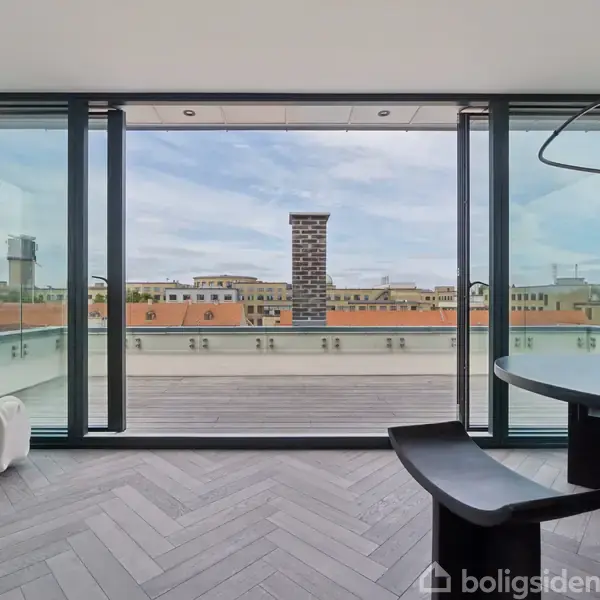 Panoramavindue åbner ud til en tagterrasse med udsigt over byens tage. Indendørs ses mørke parketgulve og en moderne lampe i loftet. Trækasse står ved glasvæggen.