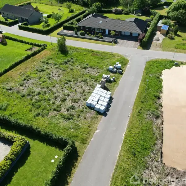 Tomt med stablede byggematerialer, omgivet af villahuse og grønne marker i et boligkvarter. Fotograferet fra oven, materiale ligger på midten af den ubenyttede, græsbevoksede grund.