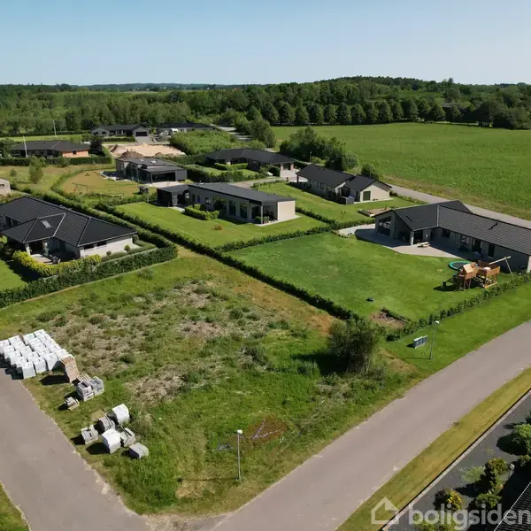 Flere moderne parcelhuse står stille langs en vej i et landligt boligkvarter. Græsplænerne er velplejede, og hele området er omgivet af grønne marker og skov i baggrunden.