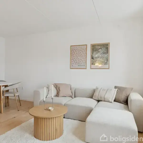 En lysegrå sofa står i en stue; ved siden af et rundt bord med stearinlys. I baggrunden ses et spisebord med fire stole. Væggene er hvide med to kunstplakater.
