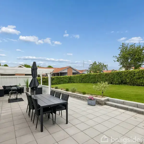 En udendørs terrasse med et sort havebord og stole, to parasoller. Stenbelægning omgiver det grønne græsareal og buske; klare, blå himmel i baggrunden.