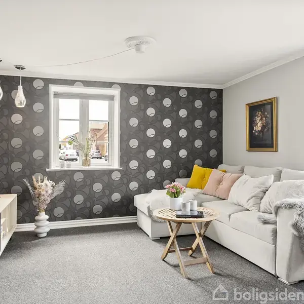 En lys stue med en hvid hjørnesofa fyldt med puder, der står mod en væg dekoreret med grå og hvide cirkler. Et rundt sofabord står foran sofaen, og et fjernsyn er placeret på et lavt skab. Maleri hænger på væggen. Lamper hænger ned fra loftet.