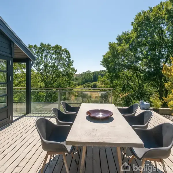 Udendørs spisebord har seks stole omkring sig, omgivet af en træterrasse. Terrassen er ved siden af et hus med glasdøre og omgivet af frodige træer og en klar himmel.