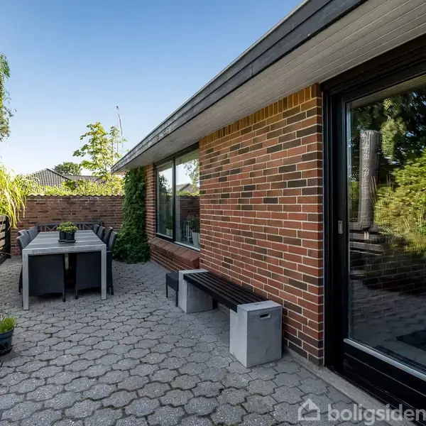 En baggård med et rektangulært bord omgivet af fire stole står på en stenbelagt terrasse. Et rødt murstenshus danner baggrund, omgivet af buske og træer.