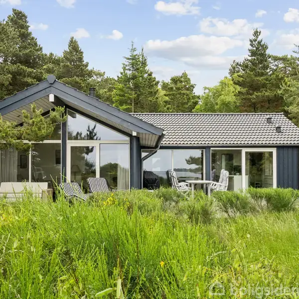 Et sortmalet sommerhus med store glasfacader står stille omgivet af tæt, grøn bevoksning. Solstole og terrassebord indikerer en udendørs terrasse, mens skoven skaber en rolig atmosfære.