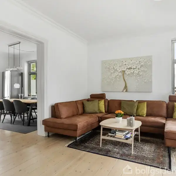 Brun lædersofa står i hjørnet af en lys stue med trægulv; på væggen hænger et maleri af et træ. I baggrunden ses en spisestue med spisebord og sorte stole.