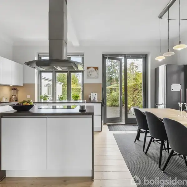 Køkken med hvidt interiør og moderne apparater, såsom emhætte over kogeø. Spisebord med sorte stole til højre. Vinduer og glasdøre med udsigt til have.