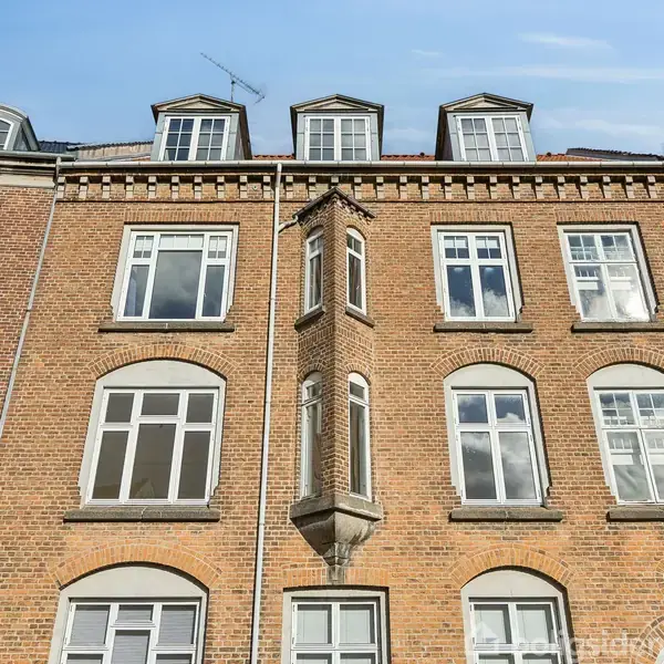 Murstensbygning med flere hvide vinduer strækker sig vertikalt mod en blå himmel. Bygningen har tre etager og en tagkonstruktion med små kviste.