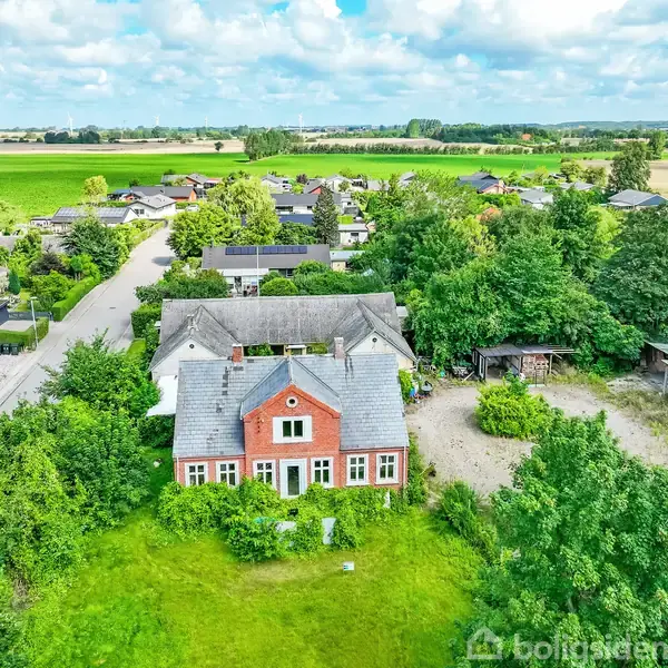 Et rødt murstenshus med gråtag, beliggende i en frodig have omkranset af træer. Omkring huset ses en række andre boliger og åbne marker under en blå himmel med skyer.