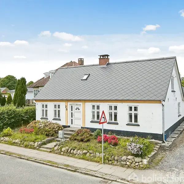 Et hvidt hus med gråt sadeltag ligger ved en rolig vej omgivet af en velplejet have med farverige blomster. Der er en vejskilt foran huset og en sti til højre.