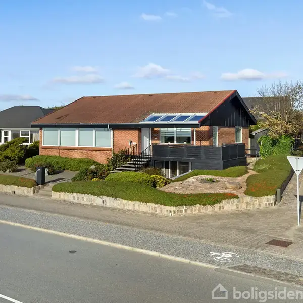 Et rødt murstenshus med skråt tag står på en hjørnegrund omgivet af lav beplantning og fortov. Det er placeret i et boligområde med asfalterede veje og andre huse i baggrunden.