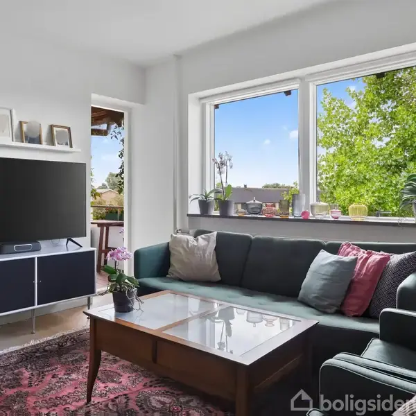 En stue med en stor grøn sofa fyldt med puder placeret ved et vindue. Foran sofaen står et sofabord, omgivet af planter og en fladskærms-TV.