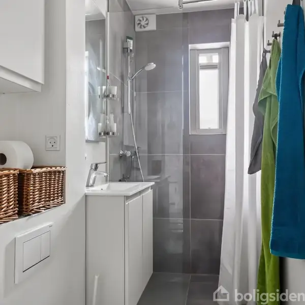 Badeværelse med bruser og gråt fliseområde, lille vindue, hvid vask på skab, kurve med toiletpapir, håndklæder hængende på kroge i hvid og grøn.