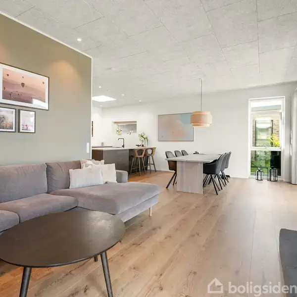 Stor stue med grå hjørnesofa, rund sort sofabord. Væggen har indrammede billeder. I baggrunden ses spisebord med sorte stole, køkkenområde og en hængende lampe.