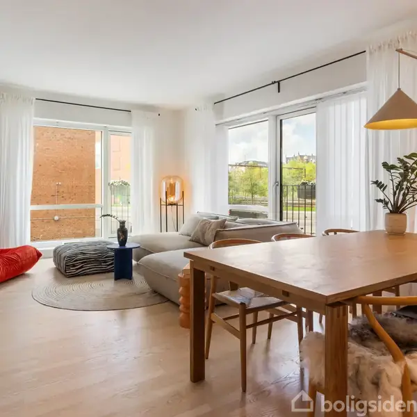 Stue med spisebord og stole, sofaer i hjørnet, og hvide gardiner ved store vinduer, der lukker lys ind. Planter og kunstværker pynter i det moderne rum.