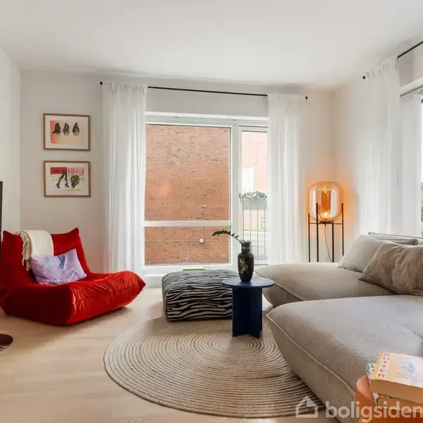 En stue med en stor grå sofa og et rundt tæppe. En rød loungestol står ved siden af et vindue med hvide gardiner. Erotisk belysning og grøn udsigt udenfor.