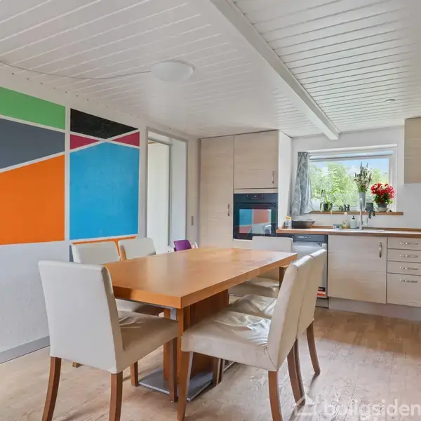 En køkken-spisestue har et bord med seks stole, ved siden af en væg dekoreret med flerfarvede geometriske figurer. Køkkenet har træskabe, et vindue og en vase med blomster.