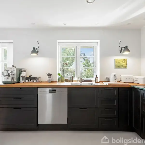 Køkken med sorte skabe står stille, mens opvaskemaskinen er i midten. Lys kommer ind gennem vinduer, og urtepotter står ved siden af kaffemaskinen på bordpladen.