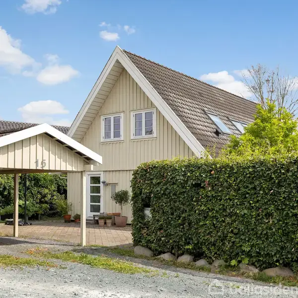 Et lyst træhus med brune tagsten og to etager står ved siden af en carport markeret med nummer "125". Omgivet af grønne buske og en grusvej under en blå himmel.