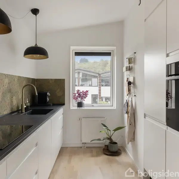 Et moderne køkken med hvide skabe og sort bordplade, hvor en indbygget ovn ses til højre. En plante og kogekande står nær et vindue med udsigt til huse.