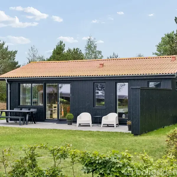 Et sortmalet træhus med rødt tegltag står i en grøn, græsbeklædt have. Foran er der en terrasse med havesæt og to hvide stole, omgivet af træer.