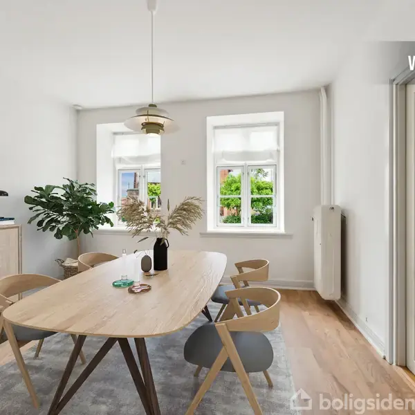 Et ovalt spisebord med stole står centralt i et lyst rum med trægulv. Der er planter på skænken, to vinduer med udsigt til grønt og teksten "VISUALISERING" i øverste højre hjørne.