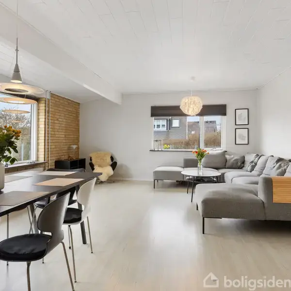 Stue med moderne sofaarrangement og spisebord, dekoreret med blomster. Store vinduer giver lys, og der er en hængende lampe over bordet. Lyse vægge og trædetaljer skaber en hyggelig atmosfære.