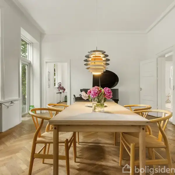 Langbord med blomsterbuketter står midt i et lyst rum. Moderne lampe hænger over bordet. Træstole omgiver bordet. Store vinduer i baggrunden slipper naturligt lys ind.