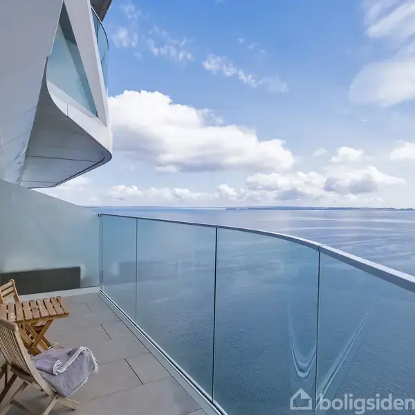 Balkon med to træstole og et tæppe placeret mod et glasrækværk, tilbyder udsigt over havet under en klar blå himmel med skyer.