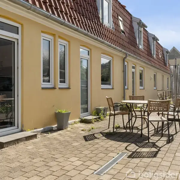 En gul bygning med rødt tegltag har døre og vinduer, der åbner ud til en brolagt gårdhave med borde og stole. Planter i potter pryder området under en blå himmel.