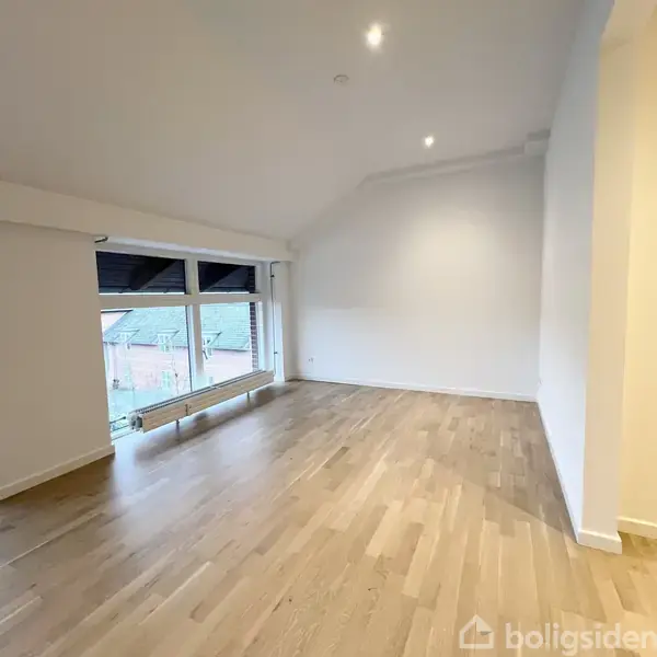 Et tomt værelse med lyst trægulv og skrå loft. En stor rude viser et udsyn til taget af omkringliggende bygninger. Vægge er hvide med en enkelt radiator under vinduet.