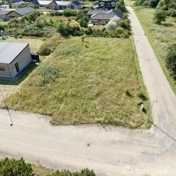 Græsbevokset tomt ved siden af et moderne hus i et boligområde. En grusvej løber langs tomtens kant, omkranset af træer og buske.
