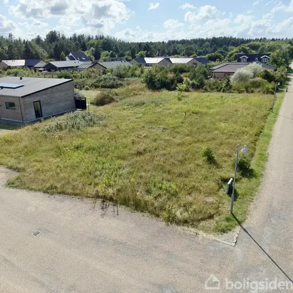 Et ubebygget græsbevokset grundstykke ligger ved siden af en moderne, enkeltstående bygning. Omgivet af en asfalteret vej, er der flere huse tæt på, og et skovklædt område i baggrunden.