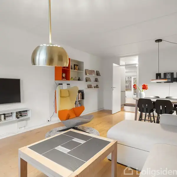 En moderne stue med en stor sofa og et sofabord, hængende lampe over bordet, fladskærms-tv på væggen; i baggrunden ses spisebord med stole og åbne døre.