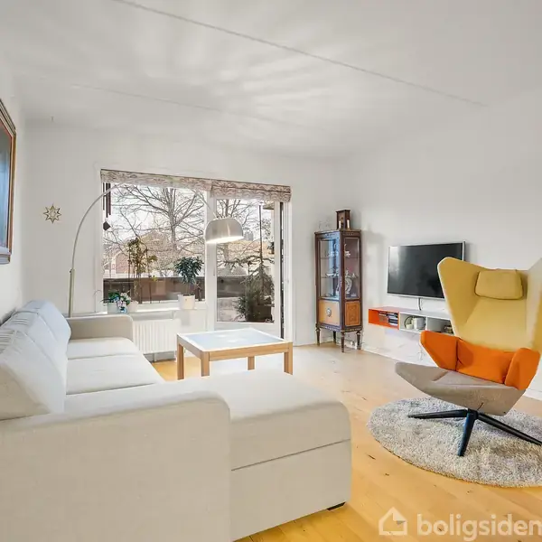 En lys stue med en hvid sofa i forgrunden, en gul-orange lænestol på et rundt tæppe og en fladskærms-TV på væggen. Store vinduer giver udsigt til en terrasse.