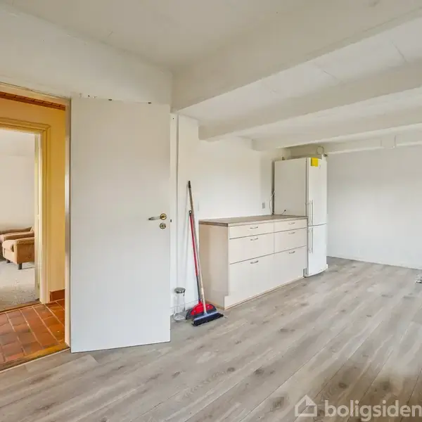 Dør åbner ind til stue med lyse vægge og trægulv, køkken på højre med skabe og køleskab, gulvskrubbe lænet mod væg; gennem døren ses en stue med sofa.